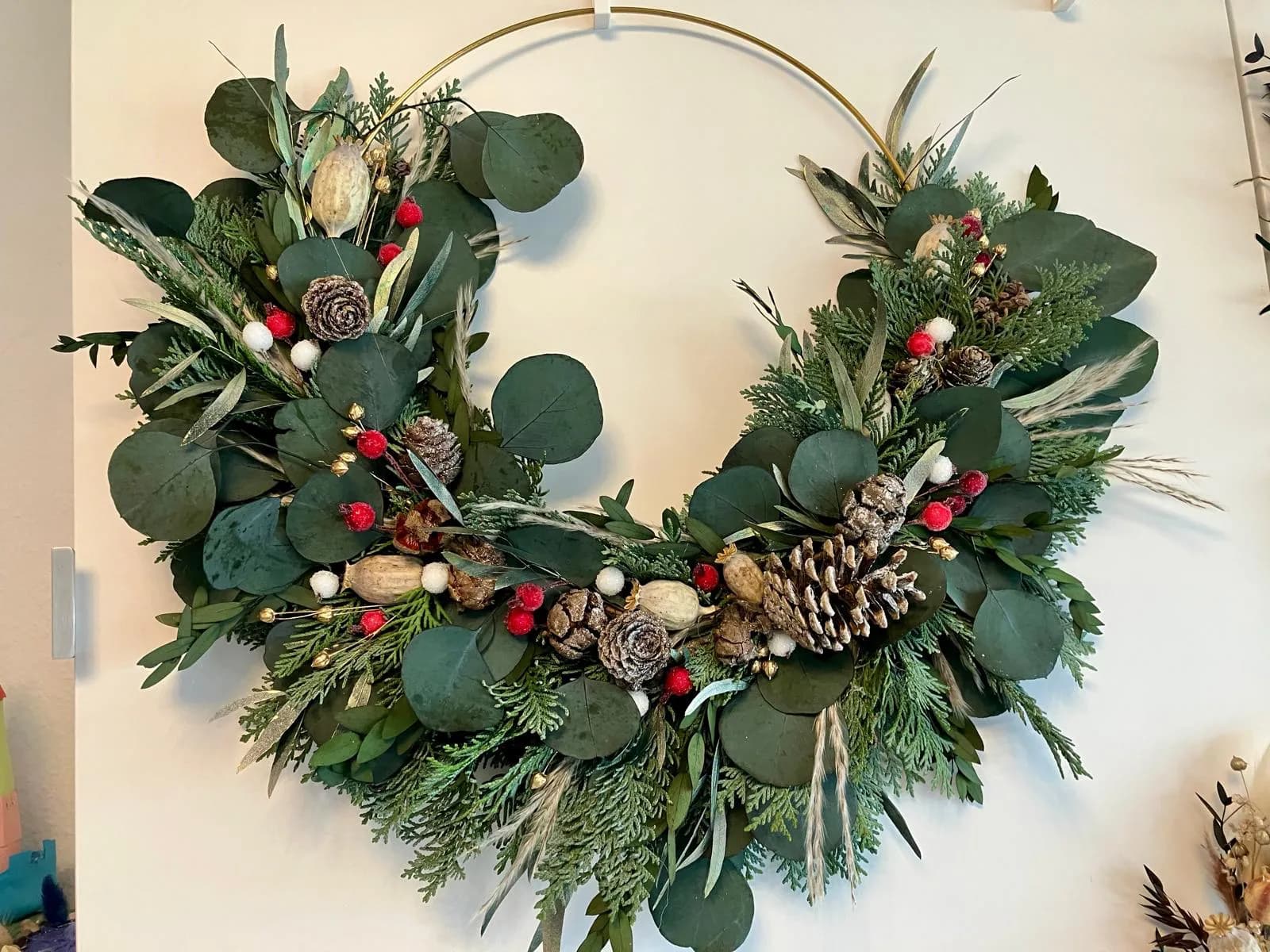 Ein halb gebundener goldener Kranzreif an der Wand, mit Eukalyptus, Thuja, verschneiten Zapfen, Mohnkapseln und roten Beeren
